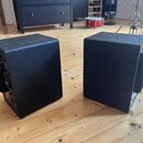 Dva černé studiové monitory v kostkovém tvaru s texturovaným povrchem. Jsou umístěny na dřevěné podlaze, zadní strana levého reproduktoru ukazuje při…