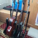 Černý stojan s čtyřmi kytarami. První kytara zleva je červená akustická kytara s vyřezem v korpusu. Další tři kytary jsou tmavé barvy, z nichž jedna…