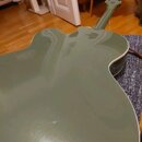 Fotografie zachycuje zadní stranu akustické kytary v olivově zelené barvě s lesklým povrchem. Nástroj má vyřez (cutaway) v těle a na hlavě jsou vidět…