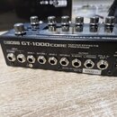 Černý kytarový efektový procesor s nápisem BOSS GT-1000CORE GUITAR EFFECTS PROCESSOR. Na zadním panelu jsou vstupy a výstupy s popisky INPUT R/MONO,…