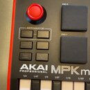 Detailní pohled na část černého MIDI kontroléru s bílými a černými klávesami. Na přístroji je vidět červený knoflík, dvě gumové pady s nápisy PAD 5 a…