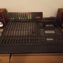 Černý digitální mixážní pult s řadou posuvníků a ovládacích knoflíků. V horní části je umístěn modul s LED indikátory a nápisem TASCAU DIGITAL STUDIO…