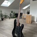 Černá elektrická kytara typu Stratocaster s hnědým hnedým hmatníkem a světlou hlavou. Na hlavě je vidět nápis Fender STRATOCASTER. Kytara stojí na če…