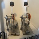 Dvě stříbrné řetězové basové pedály s černými gumovými úchyty na hrotičkách. Mechanismus pohonu je vybaven oranžovými ozubenými koly. Na obou pedálec…