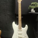 Bílá elektrická kytara typu Stratocaster s jasným dřevěným hmatníkem a hlavou. Na hlavě je viditelné logo Squier. Nástroj má tři snímače typu single-…