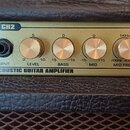 Detail ovládacího panelu zesilovače v zlaté barvě s nápisem ACOUSTIC GUITAR AMPLIFIER. Panel obsahuje dva vstupy označené jako CH1 INPUT a CH2 INPUT,…