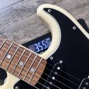 Detail elektrické kytary v krémové barvě s černým pickguardem a hnedovým hmatníkem. Na těle jsou vidět dva snímače s černými kryty. Kytara leží na di…