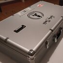 Stříbrný hliníkový kufr s kovovými hranami a rohy. Na víku jsou nalepené nálepy s nápisy „SPACESHIP IRONHEART“, „Harley Benton“ a grafickým symbolem…