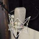 Stříbrný kondenzátorový mikrofon v šokabsorbujícím držáku. Na těle mikrofonu je viditelný nápis NEUMANN a text MADE IN GERMANY. Povrch mikrofonu i dr…