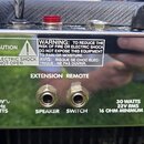 Detail zadní strany zesilovače s kovovým, zrcadlovým povrchem. Jsou zde dva konektory s popisky SPEAKER a SWITCH, nad nimi nápisy EXTENSION REMOTE. V…