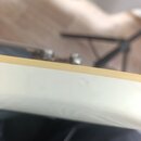 Detailní pohled na hranu krku nebo hlavy strunového nástroje v bílé barvě s kontrastním světle hnědým lemovacími pruhem. Povrch bílé lakované části v…