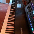Černá klávesová workstation s bílými a černými klávesami. Na pravé straně je nápis NAUTILUS MUSIC WORKSTATION a v horní části se nachází displej s ov…