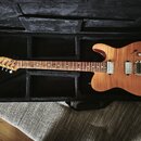 Elektrická kytara s přírodním dřevěným povrchem v černém tvrdém kufru. Nástroj má dva snímače typu single-coil, chromovaný most a ladící kolíky. Na h…