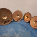 Čtyři mosazné činely různých velikostí položené na modré podlaze u bílé zdi. Každý činel má v dolní části černý nápis pAiste. Největší činel zleva má…