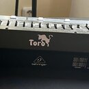 Černý MIDI kontrolér s řadou posuvníků a několika otočnými knoflíky. Na přední straně je bílý nápis „Toro“ s ikonou býka a logo „behringer“. Na zadní…