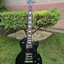 Černá elektrická kytara s typickým tvarem Les Paul, umístěná na stojanu v trávě. Na hlavě hmatníku je bílý nápis Gibson a pod ním kurzivní nápis Les…
