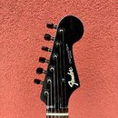 Fotografie zachycuje hlavu elektrické kytary v černé barvě. Na povrchu je bílý nápis Fender a STRATOCASTER ® s menším doplňkem MADE IN JAPAN. Nástroj…