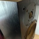 Detailní pohled na roh černého reproduktoru s povrchem imitujícím dřevěnou strukturu. Na přední straně je vidět horní středový reproduktor s černým k…