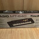 Fotografie znázorňuje kartonovou krabici s elektronickým klávesem. Na krabici je uveden nápis CASIO MT-640 TONE BANK KEYBOARD a v pravém dolním rohu…