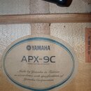 Fotografie zachycuje vnitřní štítky kytary přilepené na světlý dřevěný povrch. Oválný štítek nese logo a nápis YAMAHA, modelové označení APX-9C a tex…