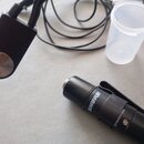 Na fotografii je černý mikrofon s připevněným klipem a připojený kabel s černým rozbočovacím prvkem. Na těle mikrofonu je bílý nápis SHURE a malý ští…