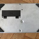 Fotografie zachycuje spodní stranu stříbrného elektronického zařízení s kovovým povrchem. V horní části je černý plastový kryt baterií s nápisy BATTE…