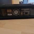 Zadní strana černého zesilovače v textilním potaху. Na přístroji jsou vidět nápisy BEHRINGER, BX3000T a webová adresa www.behringer.com. Jsou zde dva…