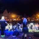 Fotografie zachycuje hudební kapelu vystupující na venkovním pódiu před davem lidí v noci. V popředí jsou ze zad vidět dva hudebníci, z nichž jeden h…