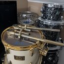 Fotografie zobrazuje několik bubnů z bicí sady. V popředí je bílý buben se zlatým hardwarem, na kterém je vidět štítek s nápisem Pearl Masters Custom…