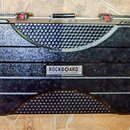 Černý transportní kufr s texturovaným povrchem a vodorovnými žebrováním. Uprostřed je umístěn kovový štítek s nápisem ROCKBOARD by WARWICK. Povrch vy…