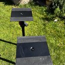 Dvě černé kovové platformy na stojanech, pravděpodobně držáky na noty nebo tablety. Povrchy jsou matně černé, přičemž spodní platforma vykazuje vidit…