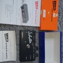 Černý efekt pedál s nápisy Schaller, TREMOLO a SPEED. Na spodní části jsou popisky AMPLITUDE, FREQUENZ, OUTPUT a INPUT. Součástí je oranžová krabice…