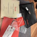 Fotografie zobrazuje soubor dokumentů a příslušenství k hudebnímu nástroji. Je zde vidět černý kožený obal s červeným logem Fender, certifikát pravos…