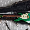 Zelená elektrická kytara s bílým pickguardem a zlatými potenciometry a hardwarem v černém tvrdém kufru. Na hmatníku je viditelné logo "Ibanez". Kytar…