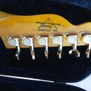 Detail hlavy elektrické kytary z jasného dřeva s osmoticým povrchem. Na dřevě je černý nápis „Fender Custom Shop“ a pod ním modré sériové číslo „SZ51…