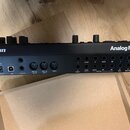 Zadní strana černého elektronického nástroje s nápisy „elektron“ a „Analog Rytm MKII“. Jsou zde vidět napájecí vypínač, konektory pro 12VDC 2A a USB,…