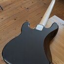 Fotografie zachycuje zadní stranu elektrické basové kytary s černým bodies a světlým dřevěným hnedem. Na kovém krytu krkovnice je vyryto logo Squier.…