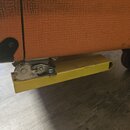 Detail spodní části oranžového komba s texturovaným povrchem. Pod korpusem je připevněna žlutá kovová lišta s mechanismem z anodizovaného hliníku. Na…