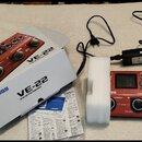 Na fotografii je červený vokální procesor BOSS VE-22 Vocal Performer v originální bílé krabici s nápisy BOSS a VE-22 Vocal Performer. Vedle krabice l…