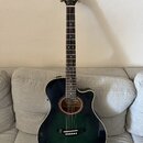 Tmavě zelená akustická kytara s cutawayem a tmavým hmatníkem. Na hlavě je bílý nápis YAMAHA a zlatá ornamentální ozdoba. Nástroj má zlaté ladičky a b…