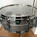 Stříbrný snare drum s chromovaným obručováním a mechanismem. Na blaně je viditelný nápis „REMO“ a „HEAVY CLEAR“. Povrch nástroje je hladký, bez vidit…