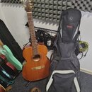 Akustická kytara s přirozeným dřevěným povrchem a hnědým krčkem. Na hlavě nástroje není žádný viditelný nápis ani logo. Vedle kytary stojí černý polo…