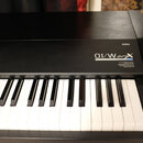 Černý digitální klavír s bílými a černými klávesami. Na horním panelu je vpravo nápis KORG a pod ním modelové označení 01/w proX MUSIC WORKSTATION. V…