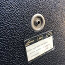 Detail černého povrchu basového komba s texturou. Na povrchu je kovový konektor a štítek s nápisy: „ELECTRONICS CORP“, logo „Peavey“ a adresa „711 A…