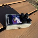 Stříbrný efekt pedál s černým ovládacím panelem a fialovým polem. Na panelu jsou nápisy electro-harmonix, BASS CLONE a popisy ovládacích prvků AMP, B…
