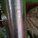 Detail stříbrného kovového trupu saxofonu s ornamentálním rytím. Na povrchu je nápis MADE BY, pod ním nečitelný podpis a text ELKHART IND. U.S.A. Pov…