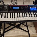 Černý digitální klavír s bílými a černými klávesami, umístěný na černém X-stojanu. Na pravé straně panelu je bílý nápis Roland Juno-DS a sekce PHRASE…