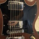 Detail elektrické kytary s hnědým dřevěným korpusem a černým pickguardem. Nástroj má dva chromované humbuckery, chromovaný kobylník a strunník. Povrc…