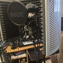 Fotografie zachycuje mikrofon v akustickém odrazovém štítu s popfiltrem. Na popfiltru i na mikrofonním krytu je viditelný nápis RØDE. V levé části sn…