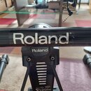 Detail černé kovové konstrukce elektronických bicích. Na vodorovné trubici je bílý nápis Roland, který vykazuje známky opotřebení a odloupení barvy.…